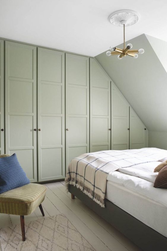 a small attic bedroom with green Pax wardrobes with dark knobs, a green bed with neutral bedding, a green chair