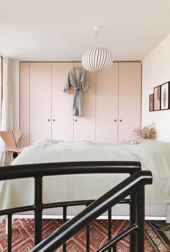 a small bedroom with built-in blush IKEA Pax wardrobes, a bed with neutral bedding, a chair and a ledge gallery wall