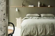 32 an olive green bedroom with paneling, a bed with light green bedding, a shelf with decor and a rattan chair
