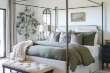 41 a farmhouse bedroom in neutrals, with a metal frame bed and olive green bedding, an upholstered bench, some art and plants