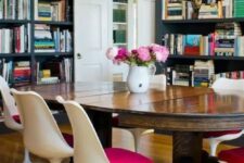a bold dining room with graphite grey walls and storage units with books, a large stained table, pink Tulip chairs and a seashell chandelier