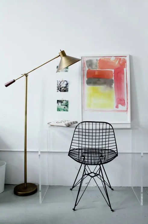 a bold space with a clear acrylic desk, a black Eames wire chair, bold artwork and a brass floor lamp is all cool