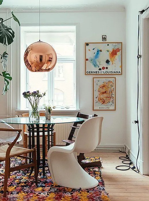a bright dining space with a glass table, mismatching chair including Panton one, bold artwork, a bright rug and a copper pendant lamp