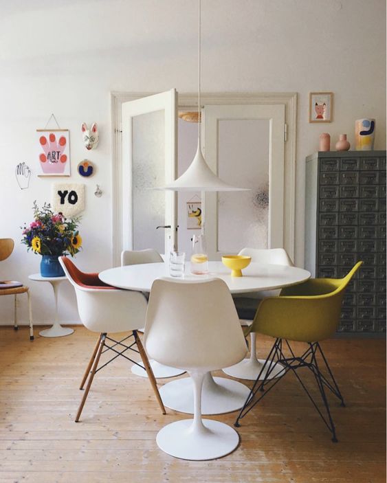 a bright dining space with a round table, Tulip and Eames chairs, a pendant lamp and some colorful home decor