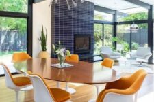 a bright mid-century modern dining room with an oval table and rust-colored tulips chairs, a fireplace clad with navy skinny tiles and a sunburst chandelier