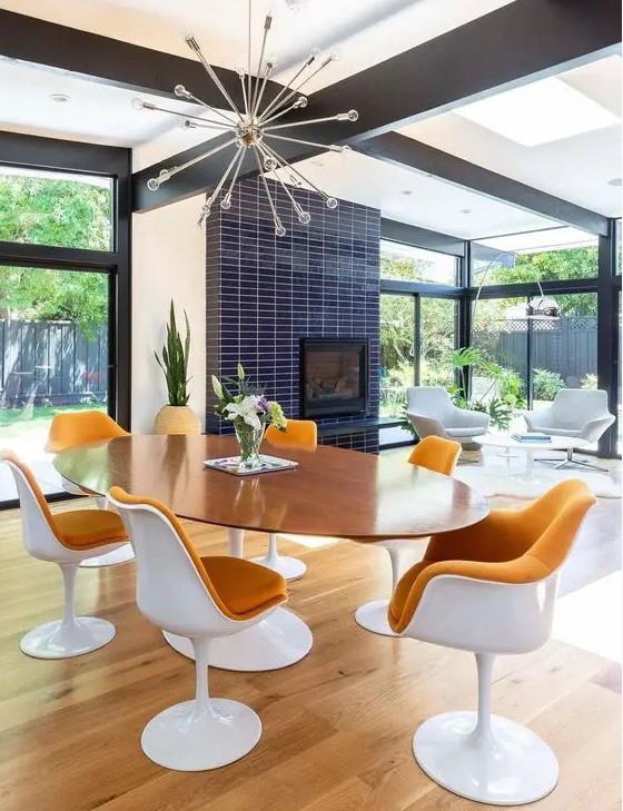 a bright mid century modern dining room with an oval table and rust colored tulips chairs, a fireplace clad with navy skinny tiles and a sunburst chandelier