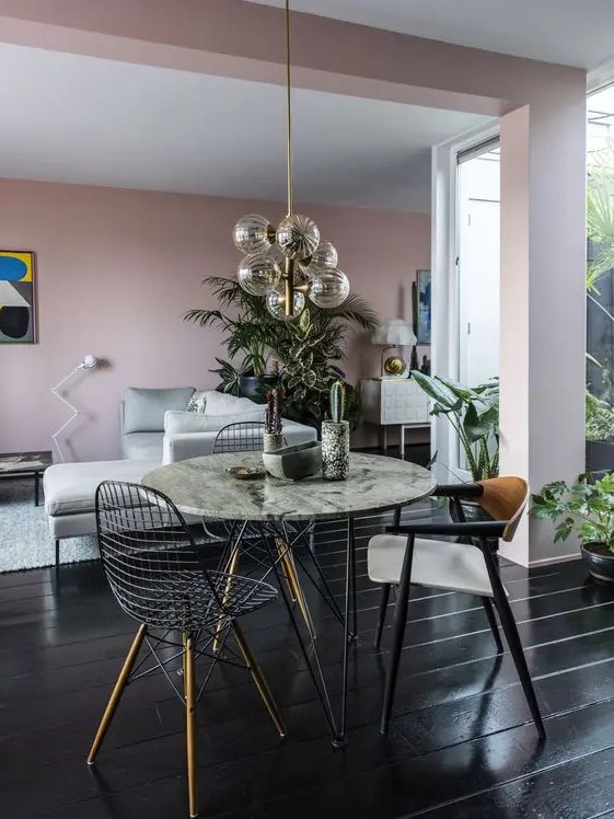 a bright space with pink walls, a grey sofa, a round table, mismatching chairs including Eames wire ones and a catchy chandelier