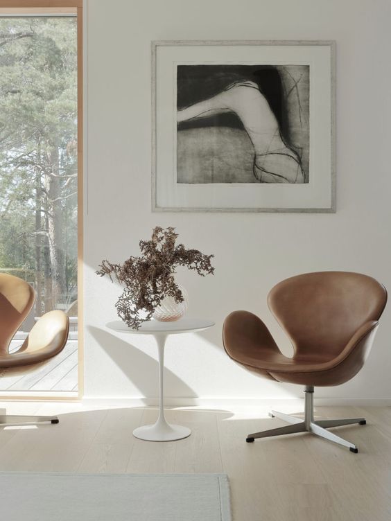 a chic nook with a couple of brown leather Swan chairs, a side table, a black and white artwork and a neutral rug