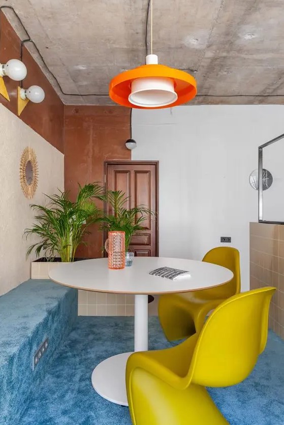 a colorful dining space with blue carpet floor that covers the bench, too, a round table, mustard Panton chairs and an orange pendant lamp