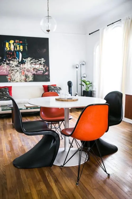 a contrasting dining room with a neutral sofa and bold pillows, an oversized artwork, a round table with Eames and Panton chairs in orange and black