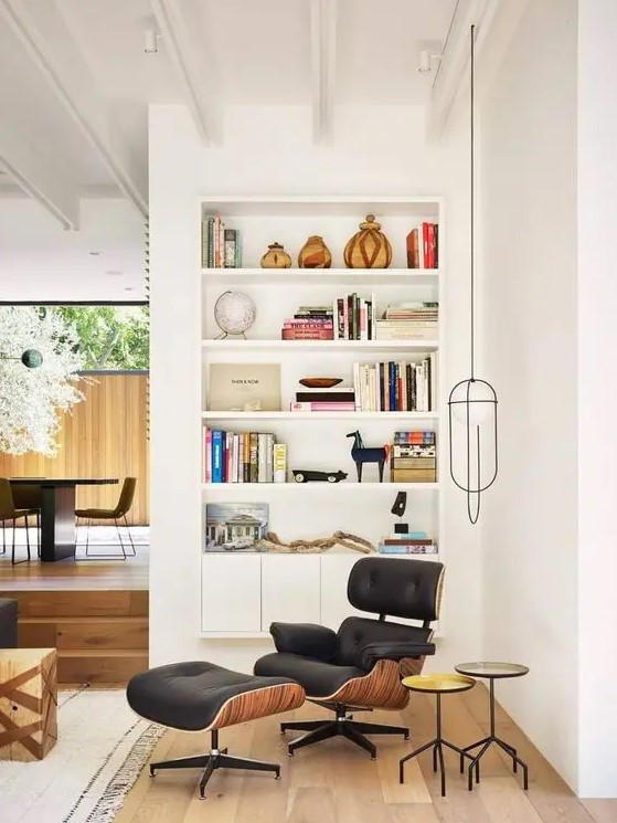 a cool nook with a floating bookcase with decor and books, a black Eames lounger and an ottoman, small side tables and a pendant lamp