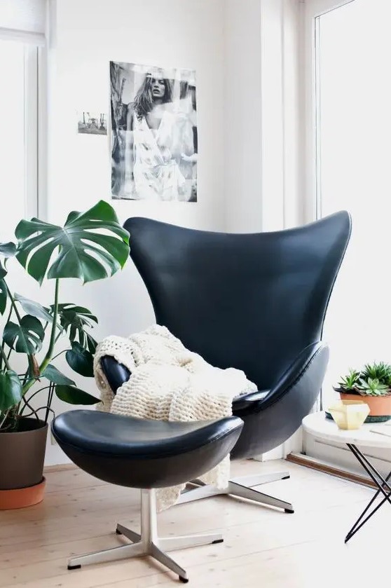 a cozy corner with a black Egg chair and a footrest, white blanket, a potted plant, some art and plants on the table