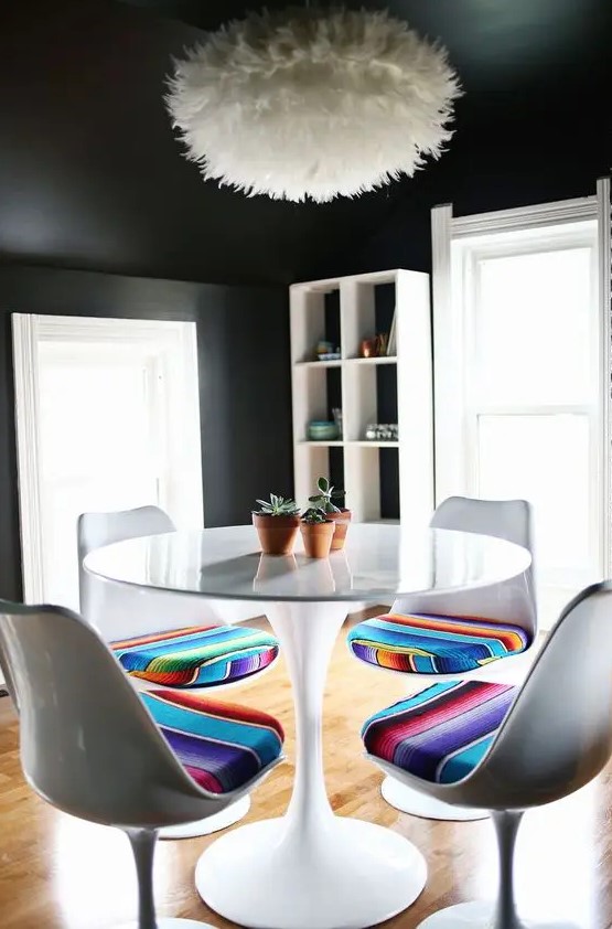 a creative dining room with black walls and a ceiling, a white storage unit, a round table, white Tulip chairs with bold upholstery