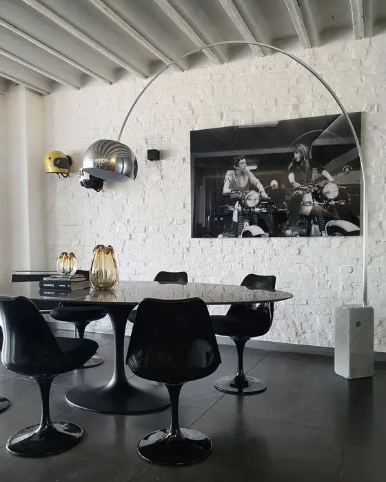 a jaw dropping dining space with a white brick wall, an oval black table, black Tulip chairs, a floor lamp and a large photo