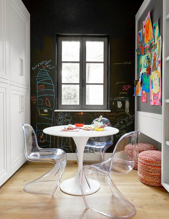 a kids' space with a chalkboard wall and a memo board, a round table and clear Panton chairs plus bright poufs