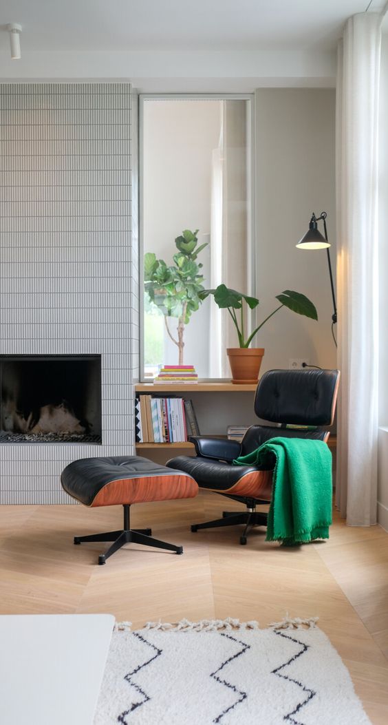 a mid-century modern living room with a fireplace clad with tiles, a black Eames lounger with an ottoman, a shelving unit and potted plants