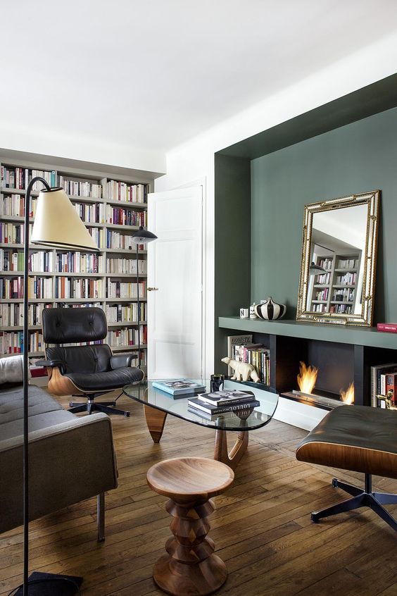 a mid-century modern living room with a green accent wall with a fireplace, bookcases, black Eames loungers, a grey sofa, coffee tables