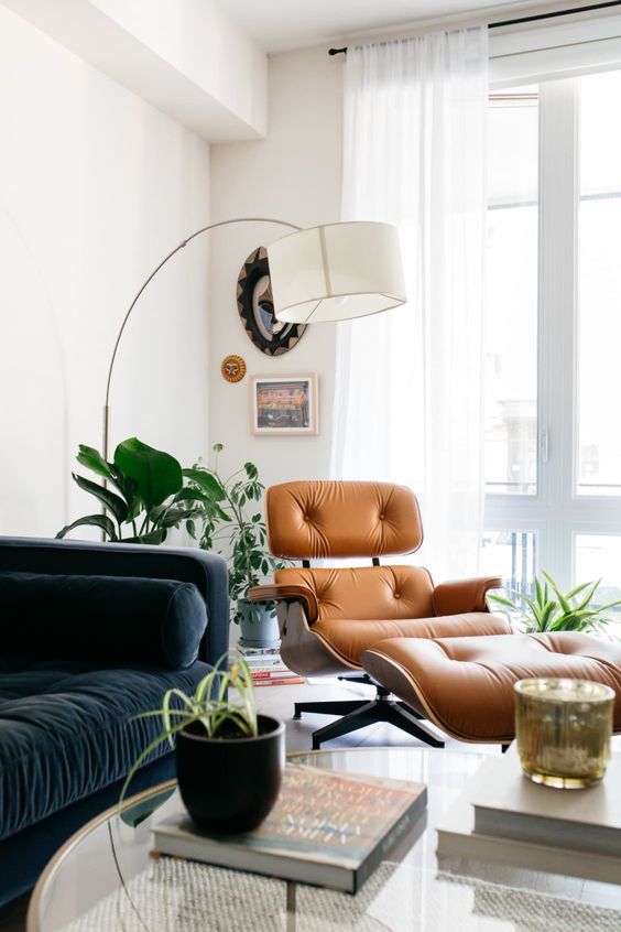 a mid century modern living room with a navy sofa, an amber leather Eames lounger, a floor lamp, some potted plants and lovely decor