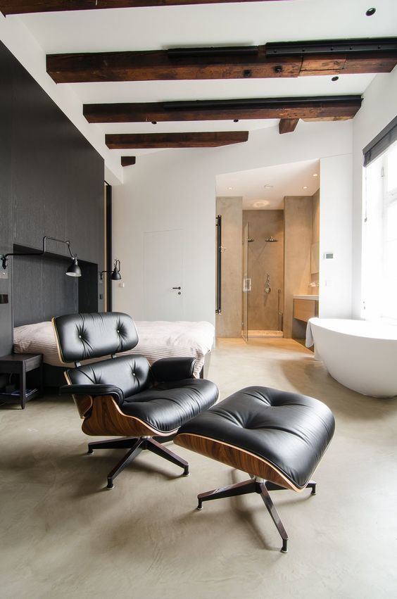 a modern bedroom with a large stained unit, a bed with neutral bedding, an oval tub, a black Eames lounger and wooden beams