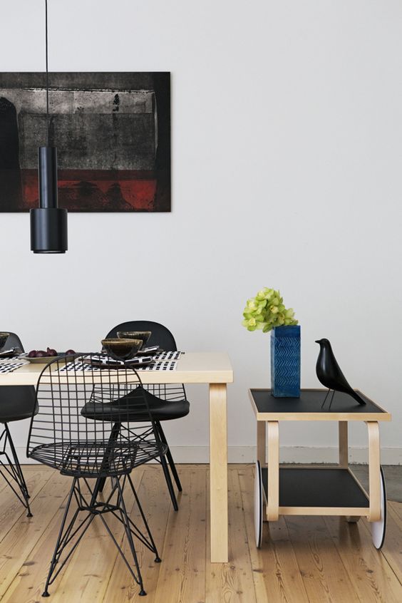 a modern dining space with a light-stained dining table, black Eames wire chairs with cushions and black pendant lamps plus some decor