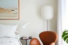 a neutral bedroom with a bed with neutral bedding, a black nightstand with a lamp, a floor lamp and an amber leather Swan chair