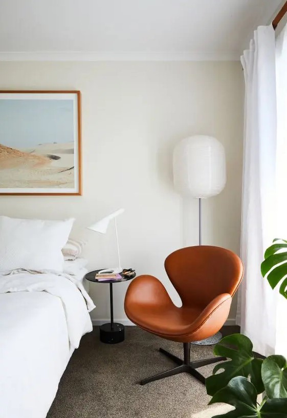 a neutral bedroom with a bed with neutral bedding, a black nightstand with a lamp, a floor lamp and an amber leather Swan chair