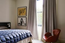 a neutral bedroom with a black bed, blue bedding, neutral curtains, bold artwork and a red Swan chair