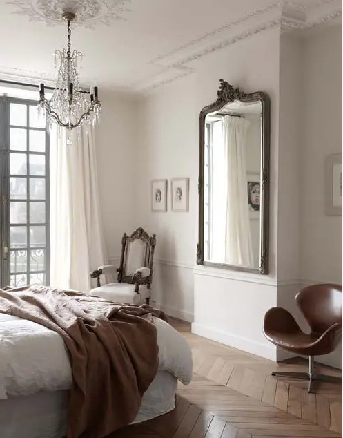 a refined Parisian bedroom with a bed and neutral bedding, a refined white chair and a brown leather Swan chair, a mirror and a crystal chandelier