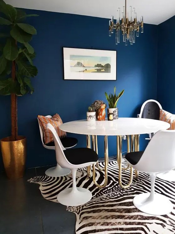 a refined and bold dining room with navy walls, a table with gold piping legs, black Tulip chairs, a zebra print rug, a cool chandelier