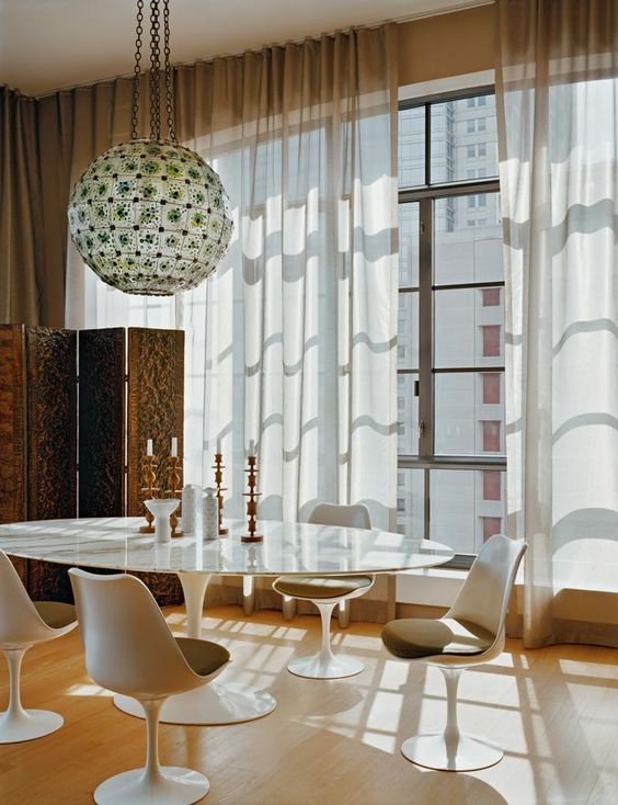 a sophisticated dining room with an oval table, Tulip chairs, a mosaic pendant lamp and a glazed wall with curtains