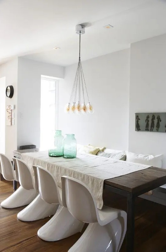 a stylish dining room with a stained table and bench, with white Panton chairs, a chandelier and some decor