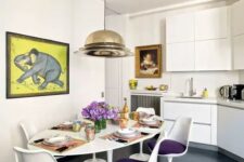 a stylish kitchen with a dining space with sleek white cabinets, an oval table with purple Tulip chairs, a cool pendant lamp and a bold artwork
