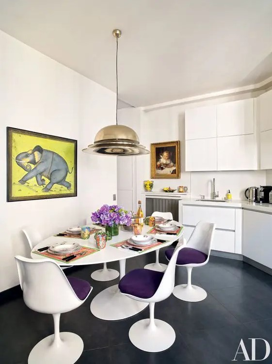 a stylish kitchen with a dining space with sleek white cabinets, an oval table with purple Tulip chairs, a cool pendant lamp and a bold artwork