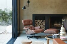 a stylish living room with a fireplace and a firewood niche, a brown Eames lounger with an ottoman, a glass coffee table and cork stools