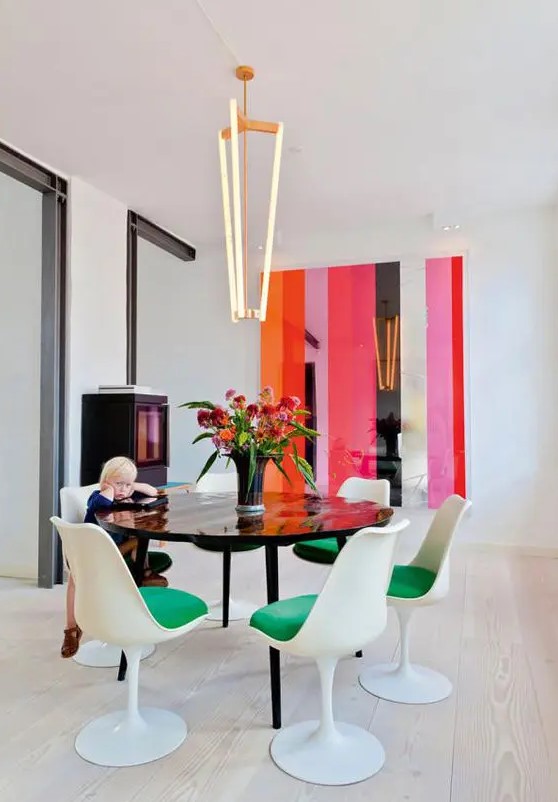 a super bright dining room with bold stripes on the wall, a storage unit, a black table and green Tulip chairs plus a gorgeous modern chandelier