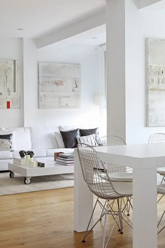 a white dining and living room with a white sofa and dark pillows, a low coffee table on casters, a white dining table and Eames wire chairs