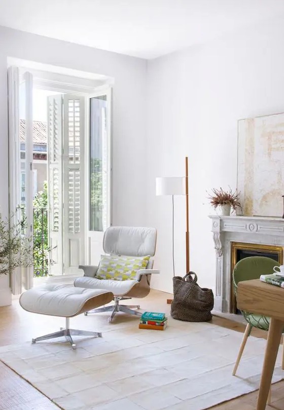 an airy white living room with a firpelace, a white Eames lounger and an ottoman, a floor lamp and a large artwork