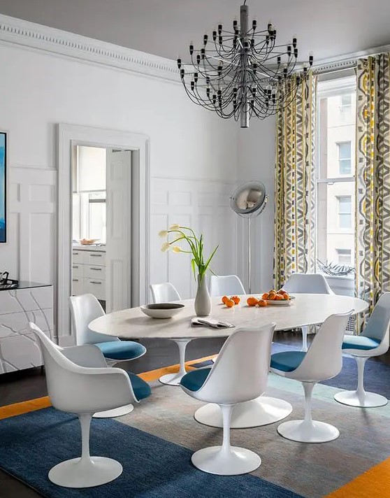 an elegant dining room with white walls, an oval table, blue Tulip chairs, a catchy chandelier and printed curtains