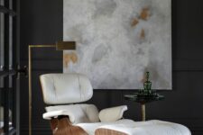 an elegant nook with black paneled walls, a creamy Eames lounger, a chic brass floor lamp and an artwork and a rug