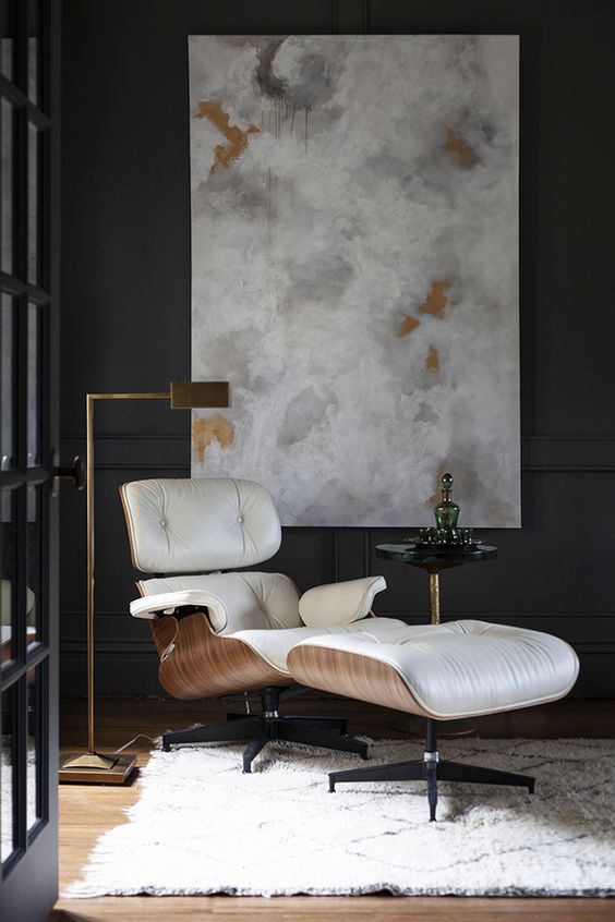 an elegant nook with black paneled walls, a creamy Eames lounger, a chic brass floor lamp and an artwork and a rug
