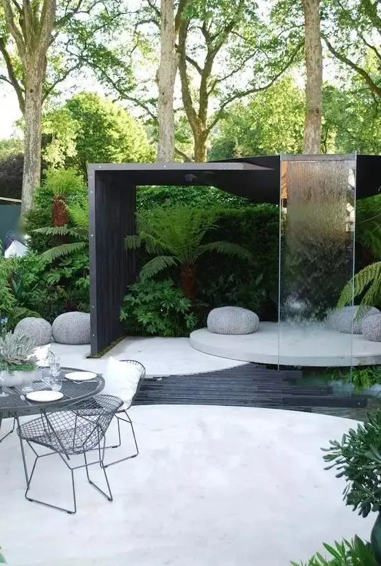 a contemporary outdoor space with a concrete round deck, a dining space, a round platform with pebbles and a waterfall of glass