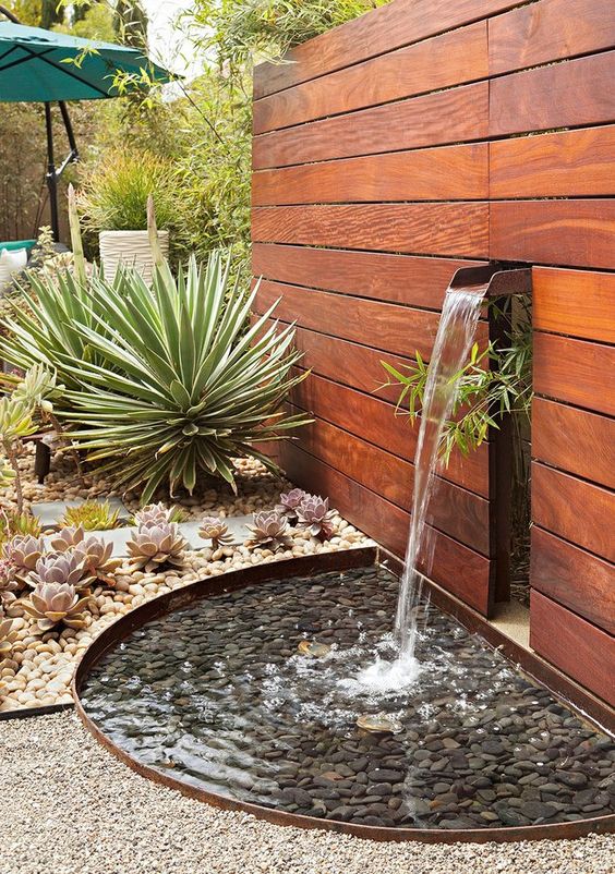 a cool desert garden with a wooden wall and a pond with pebbles on the ground, with succulents and agaves around