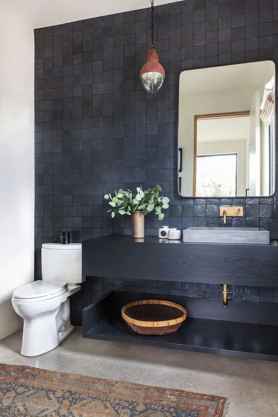 a modern bathroom with grey and black zellige tiles, a black floating vanity and a shelf, a mirror, a catchy pendant lamp and some decor