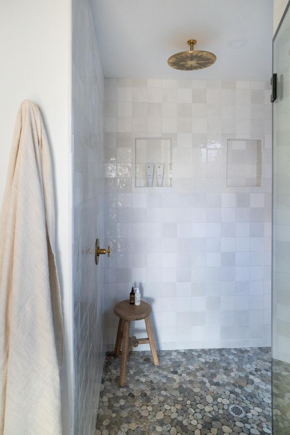 a pretty bathroom with a pebble floor and white Zellige tiles cladding the walls, brass fixtures and a wooden stool