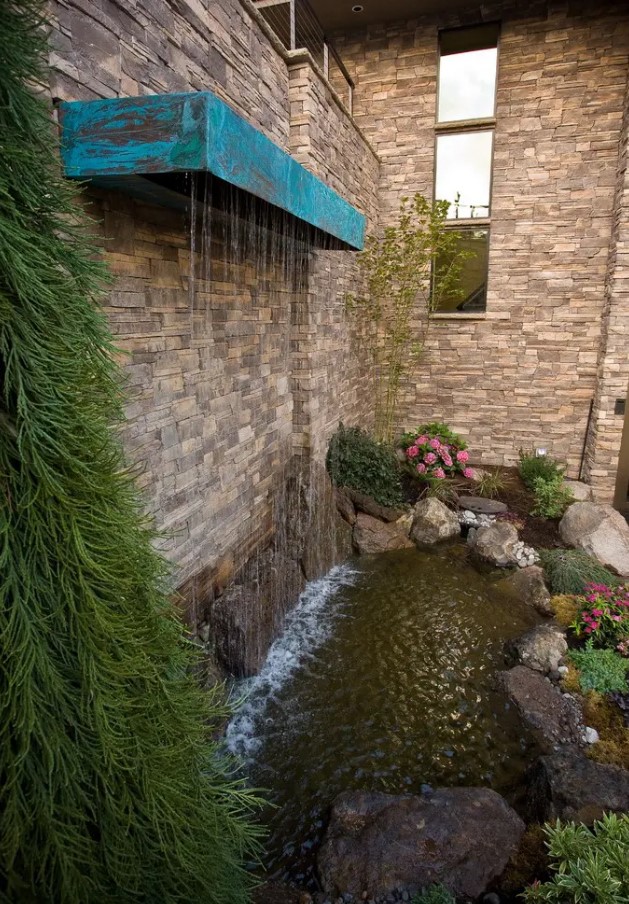 a unique waterfall of a patina piece attached to the wall, with a water feature surrounded with rocks below and some blooms