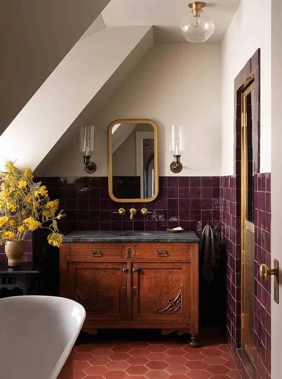 a vintage attic bathroom with a terracotta tile floor, burgundy square tiles on the walls, a stained cabinet and brass touches
