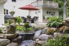 22 a beautiful and natural backyard with a dining set with a red umbrella, a grill, a ntural waterfall with large rocks and potted greenery and blooms