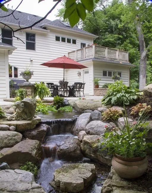 a beautiful and natural backyard with a dining set with a red umbrella, a grill, a ntural waterfall with large rocks and potted greenery and blooms