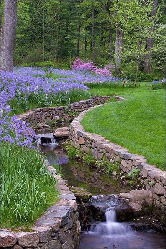 a bold garden with a green lawn, purple and pink blooms and a waterfall clad with stone and bricks is wow