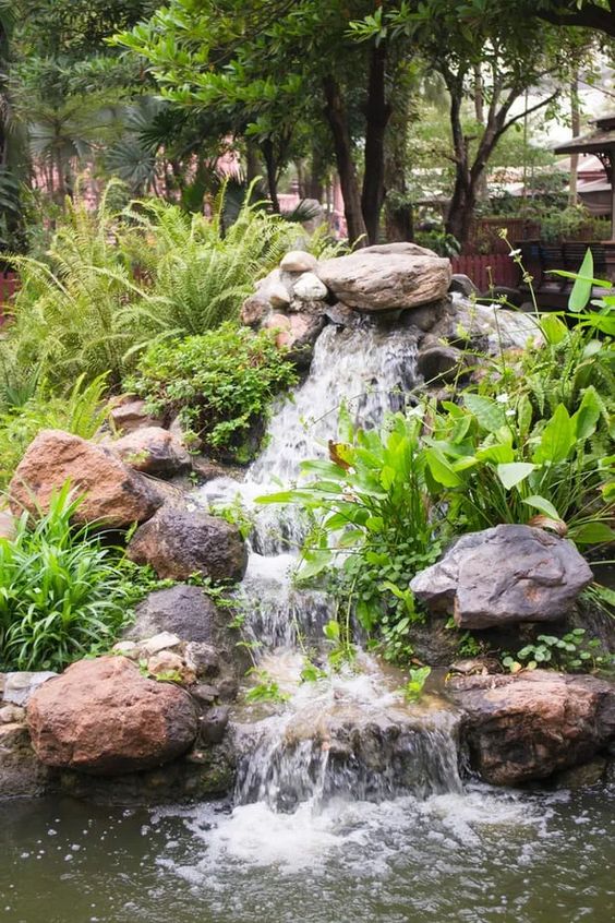 a cool backyard waterfall composed of large rocks placed on a slope, with greenery and bushes around is a lovely idea